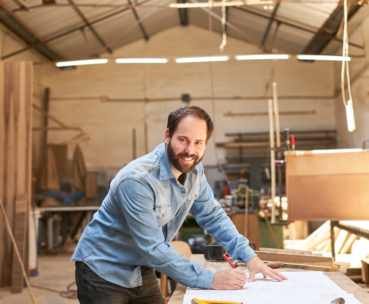 Ein Handwerker in einer Holzwerkstatt beugt sich lächelnd über seine Pläne.